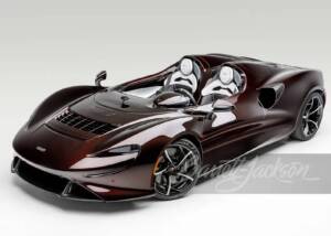 A sleek, dark metallic cherry McLaren Elva super car with white racing seats, featured on a clean white background with the Barrett-Jackson logo.