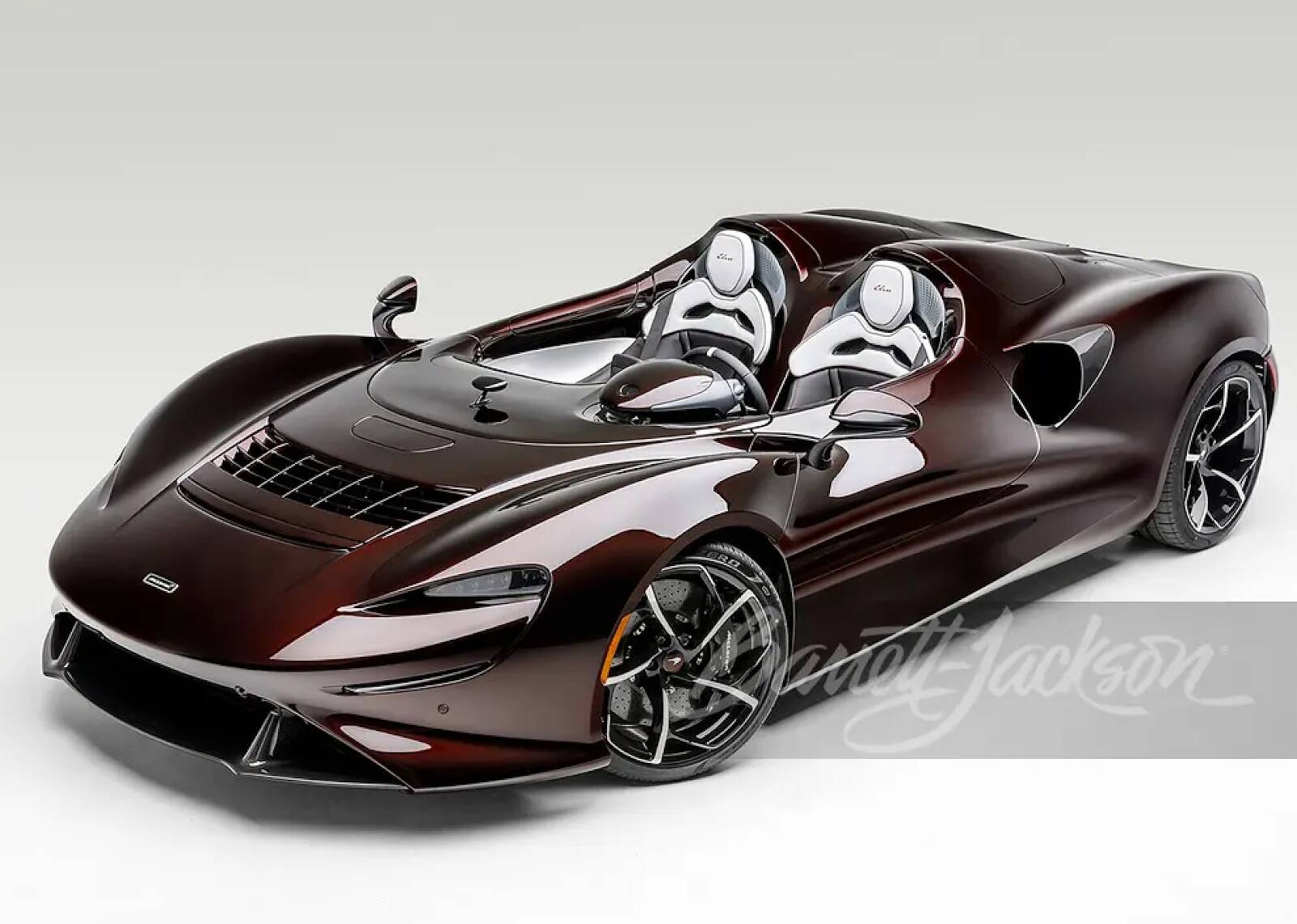 A sleek, dark metallic cherry McLaren Elva super car with white racing seats, featured on a clean white background with the Barrett-Jackson logo.
