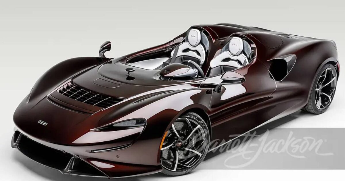 A sleek, dark metallic cherry McLaren Elva super car with white racing seats, featured on a clean white background with the Barrett-Jackson logo.